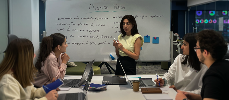 Քննարկում սպիտակ գրատախտակի մոտ, կենտրոնում սև մազերով կին